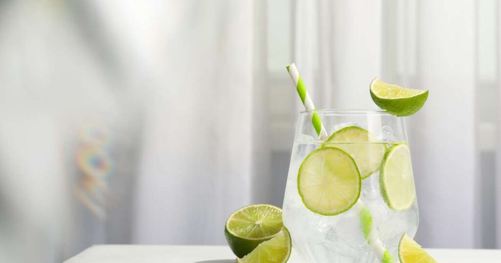 Um copo de água com limão fresco ao lado de fatias de limão e folhas verdes