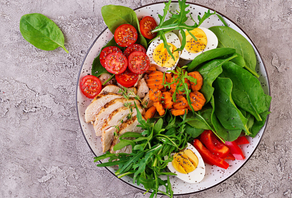 Prato colorido com legumes variados como abobrinha, pepino, alface e brócolis, acompanhados de frutas como morango e melancia, e uma xícara de chá verde.